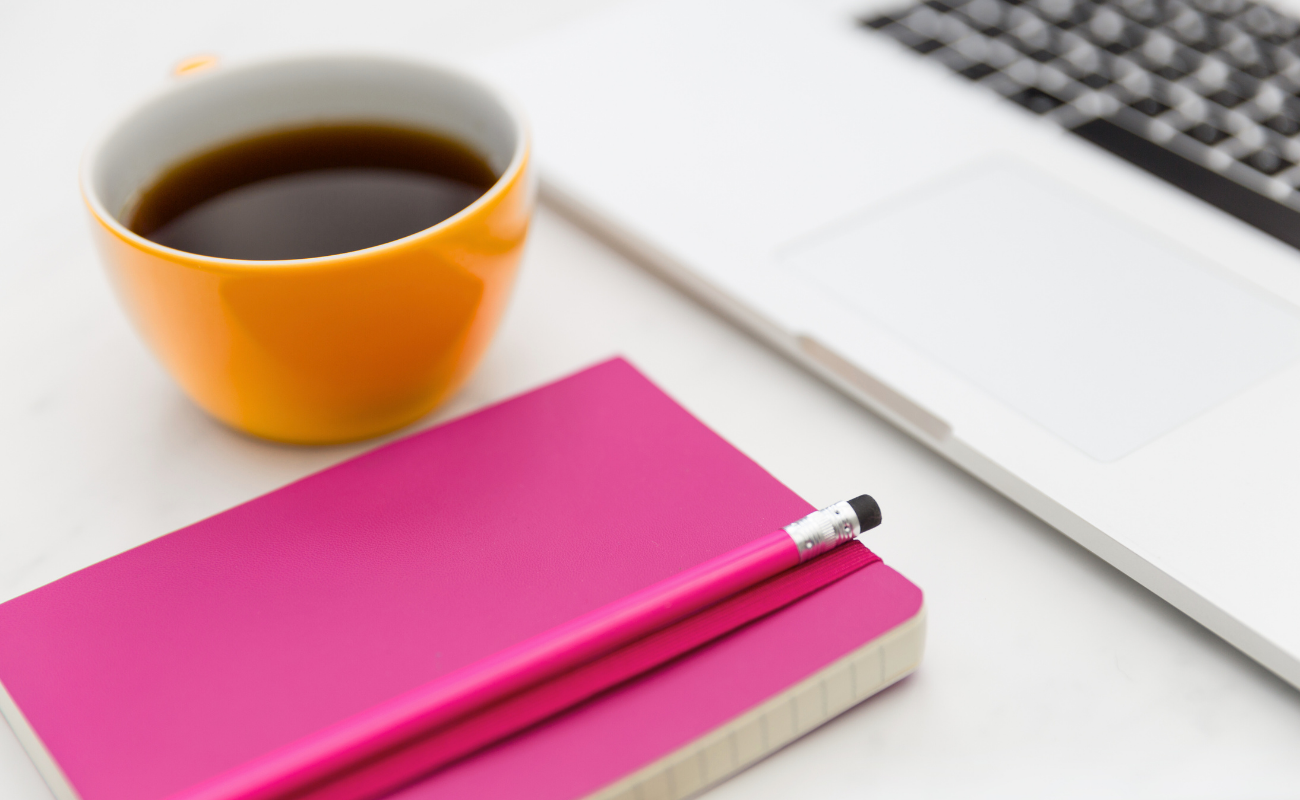 Strategic brand positioning and messaging services that define identity and drive engagement. Workspace flat lay with a coffee cup, pink notebook, and laptop, representing brand positioning and messaging strategies that define market edge and build trust.