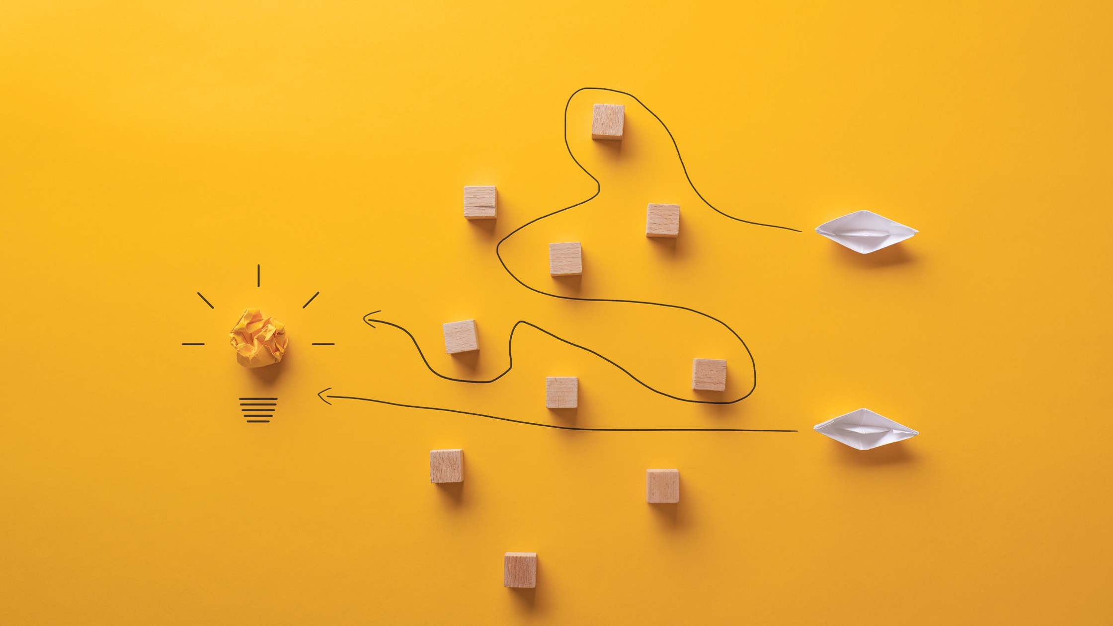 magtyne_blog image_lightbulb_paper boats_blocks A crumpled paper ball styled as a light bulb with a winding line connecting scattered wooden blocks and paper boats on a yellow background.